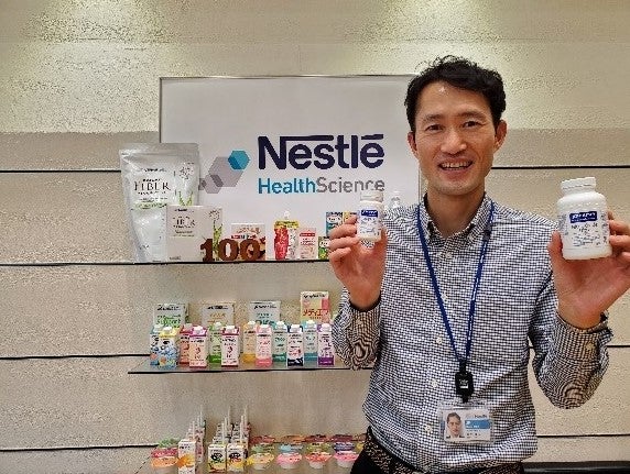 展示棚の前に立つ男性が、栄養補助食品のボトルと小箱を手に持って笑顔で紹介している。背後には健康食品ブランドのロゴが見える。