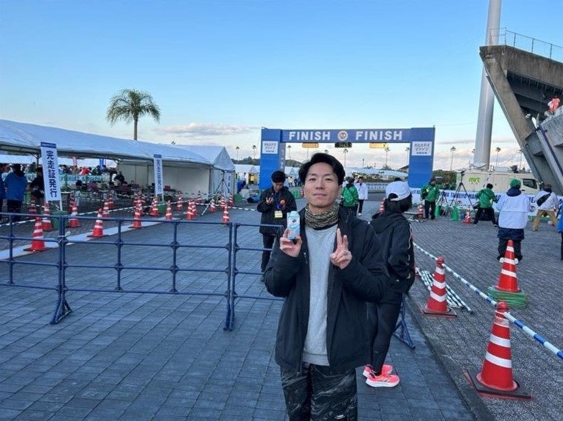 屋外イベント会場のゴールゲート前で、男性が飲料を手に持ち、ピースサインをして立っている。周囲には参加者やコーンが並んでいる。